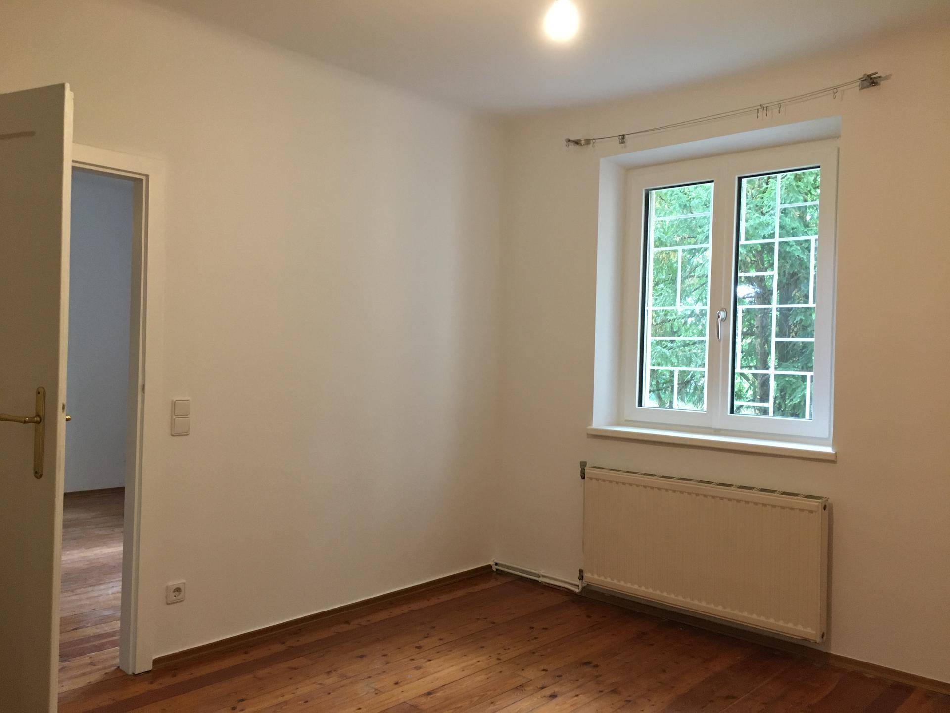 Unmöbliertes Zimmer mit Holzdielenboden, weiß gestrichenen Wänden und Fenster mit Grünblick.