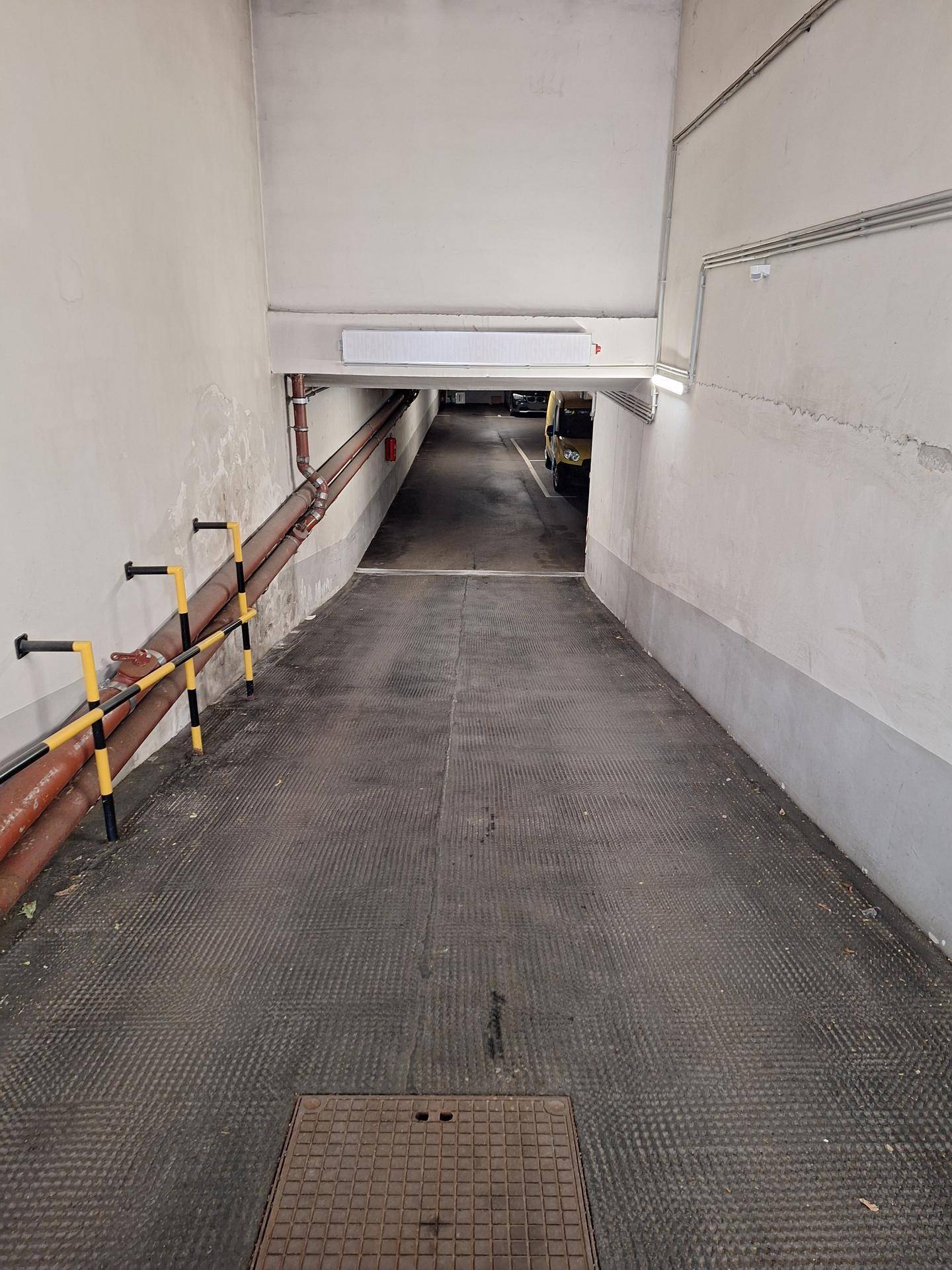 Absteigende Garagenrampe mit sichtbaren Rohren, Sicherheitsgeländern und dunklem Boden.