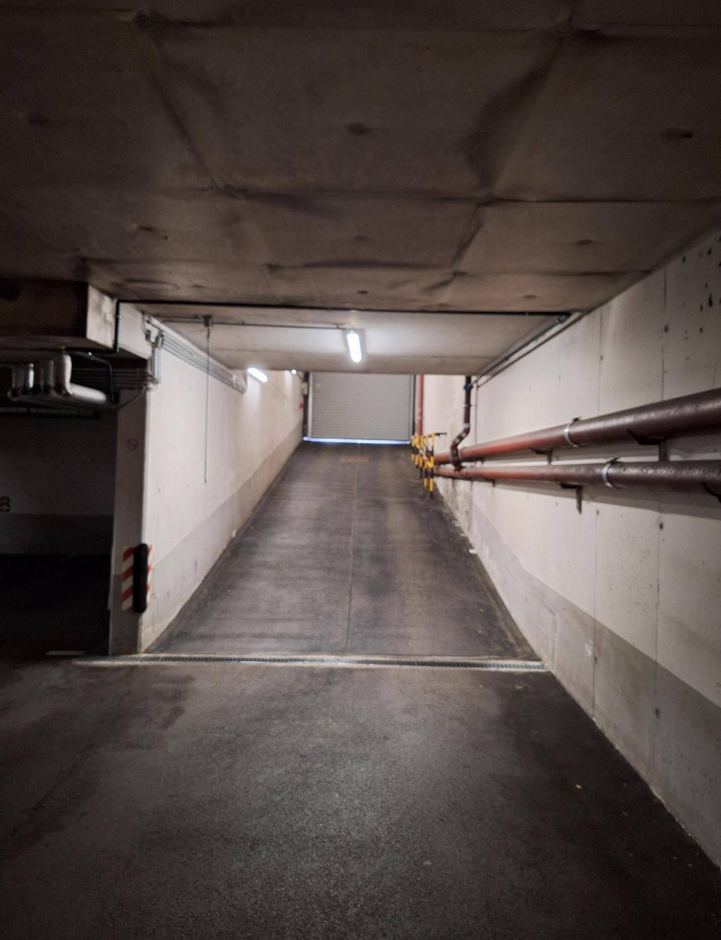 Tiefgaragenrampe führt nach unten, mit Betonwänden, Decken und sichtbaren Installationen.