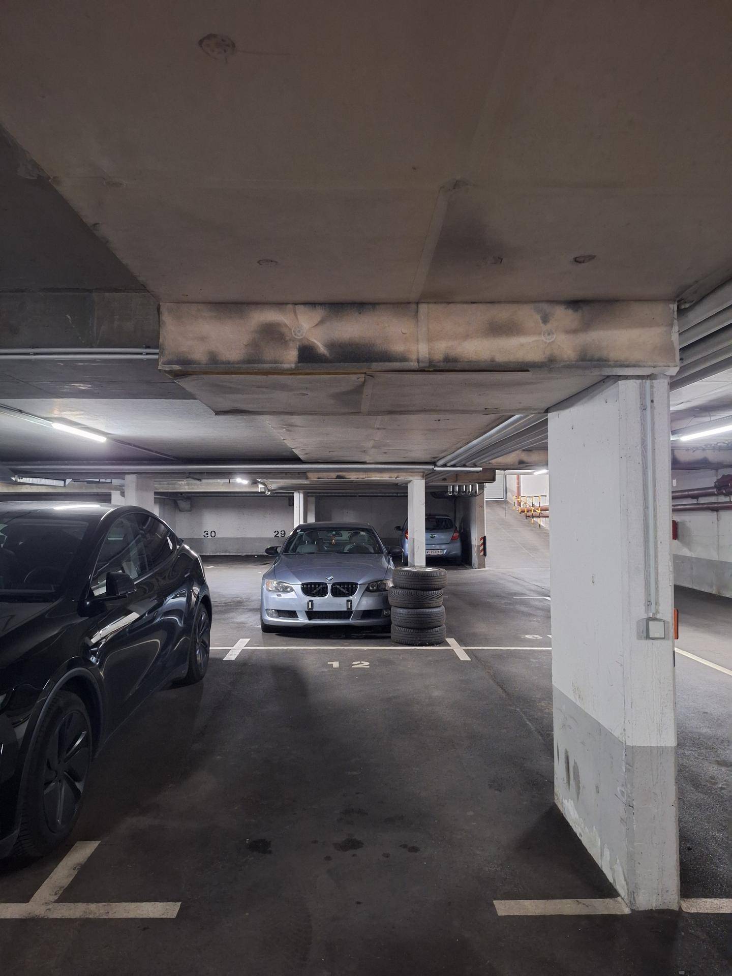 Tiefgarage mit geparkten Fahrzeugen, Betonstrukturen und sichtbaren Rohren.