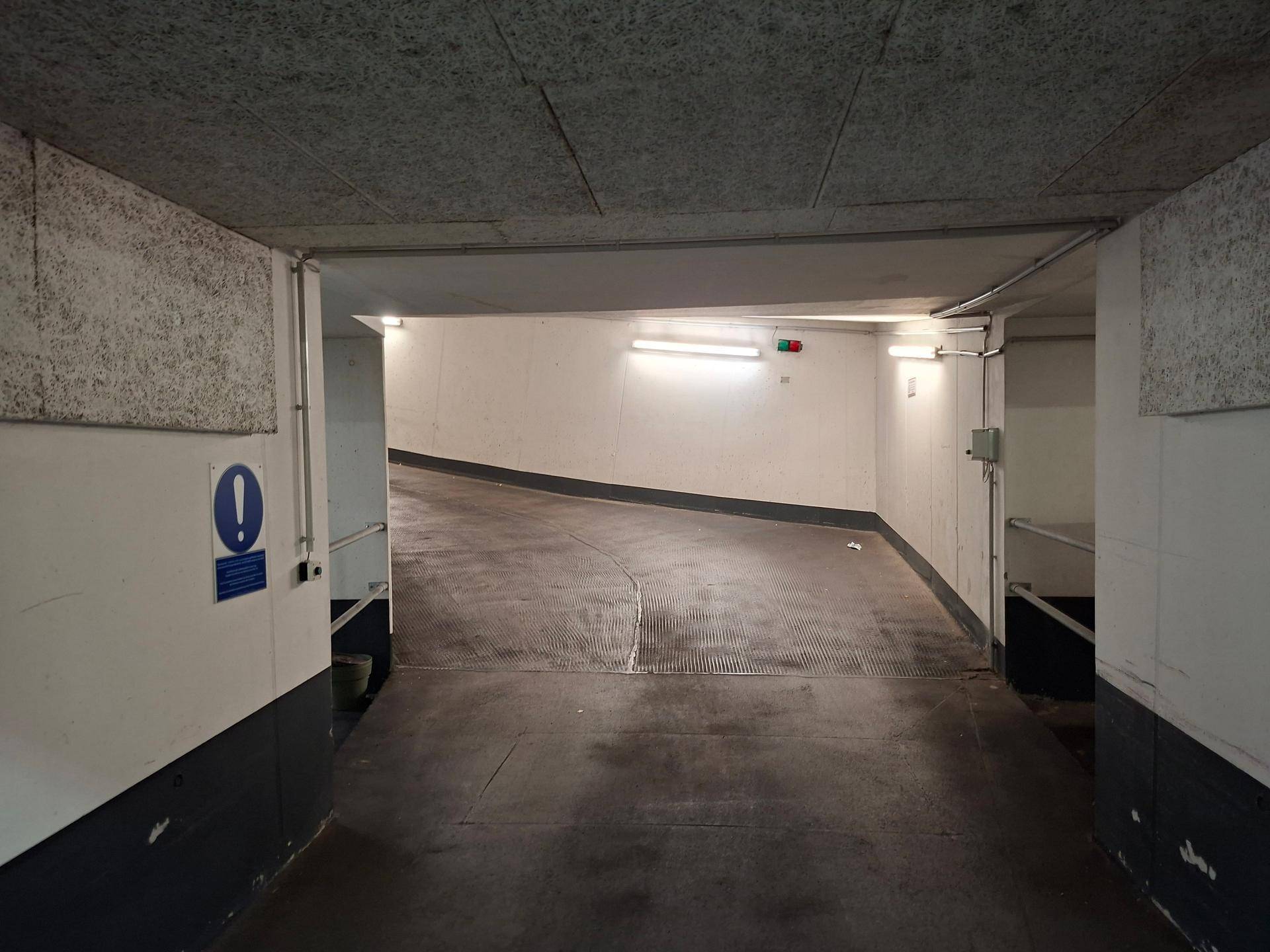 Einfahrtsrampe einer Tiefgarage mit Hinweisschild und strukturierten Wänden.