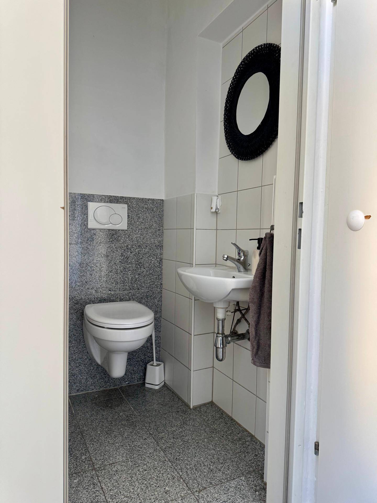 Gäste-WC mit wandhängender Toilette, kleinem Waschbecken und grauen Fliesen an Boden und Wand.