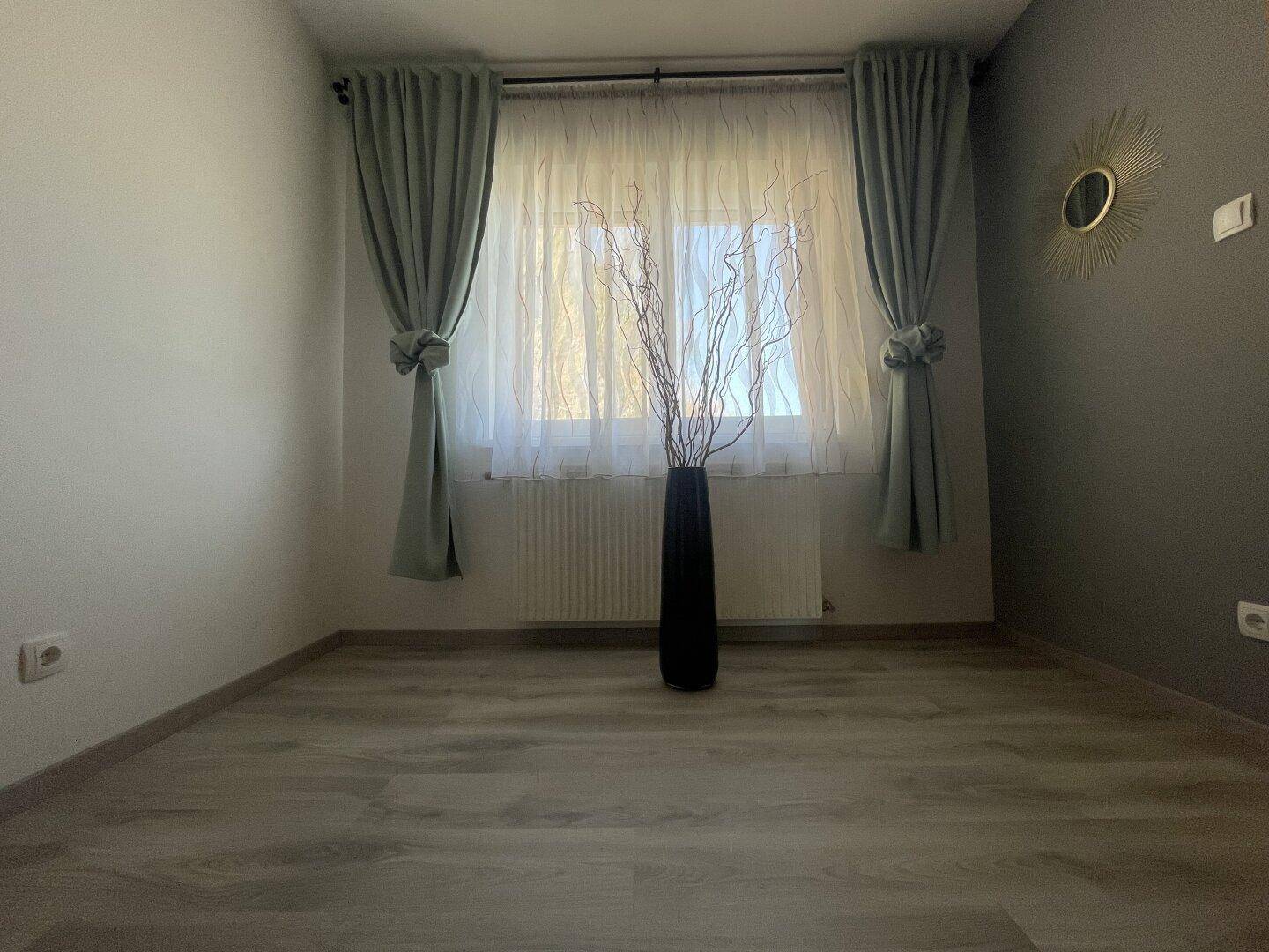 Helles Zimmer mit Laminatboden, großen Fenstern und Vorhängen, bereit zur Einrichtung.