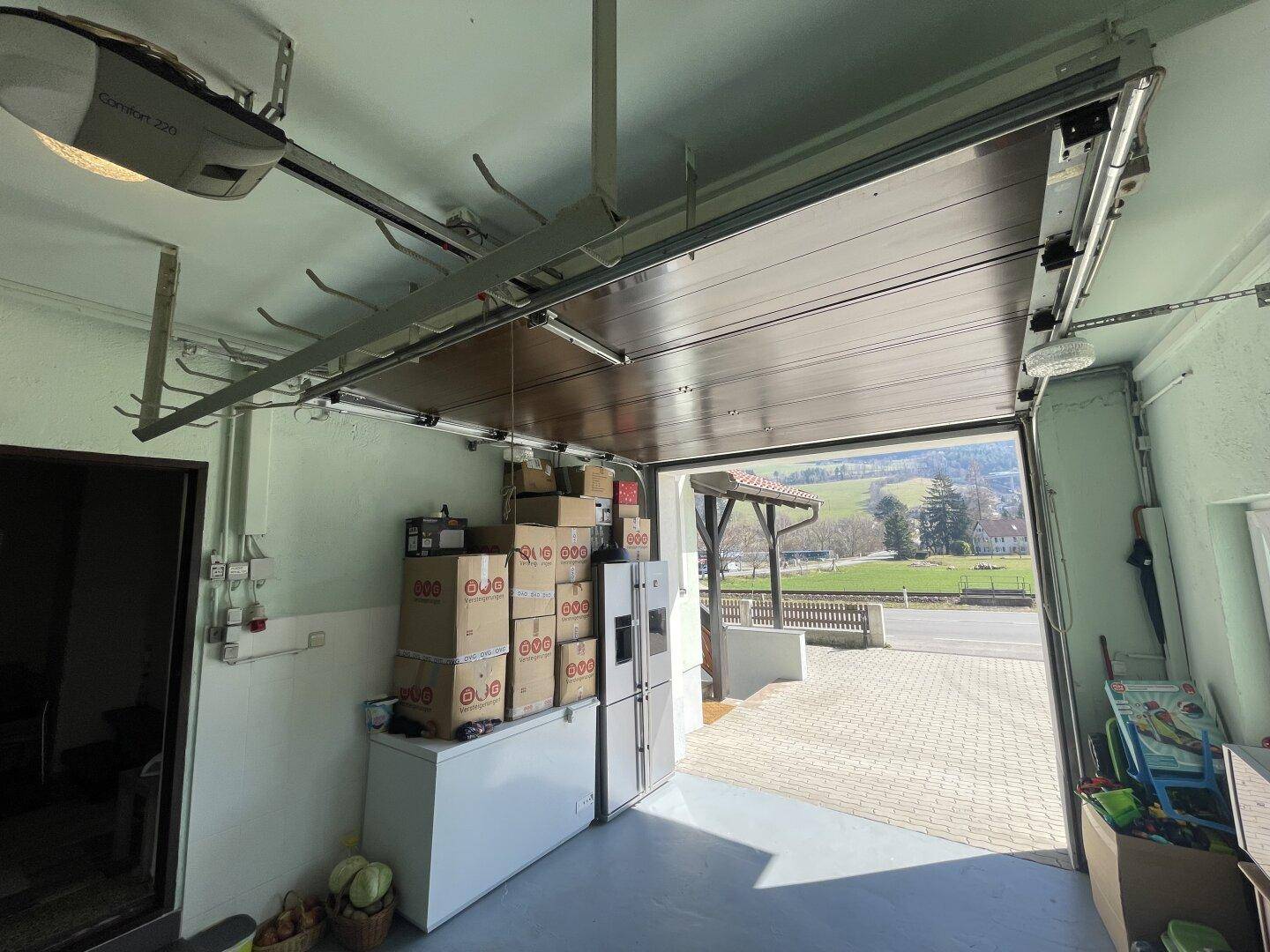Offene Garage mit Lagerflächen und Blick auf die gepflasterte Einfahrt und Umgebung.