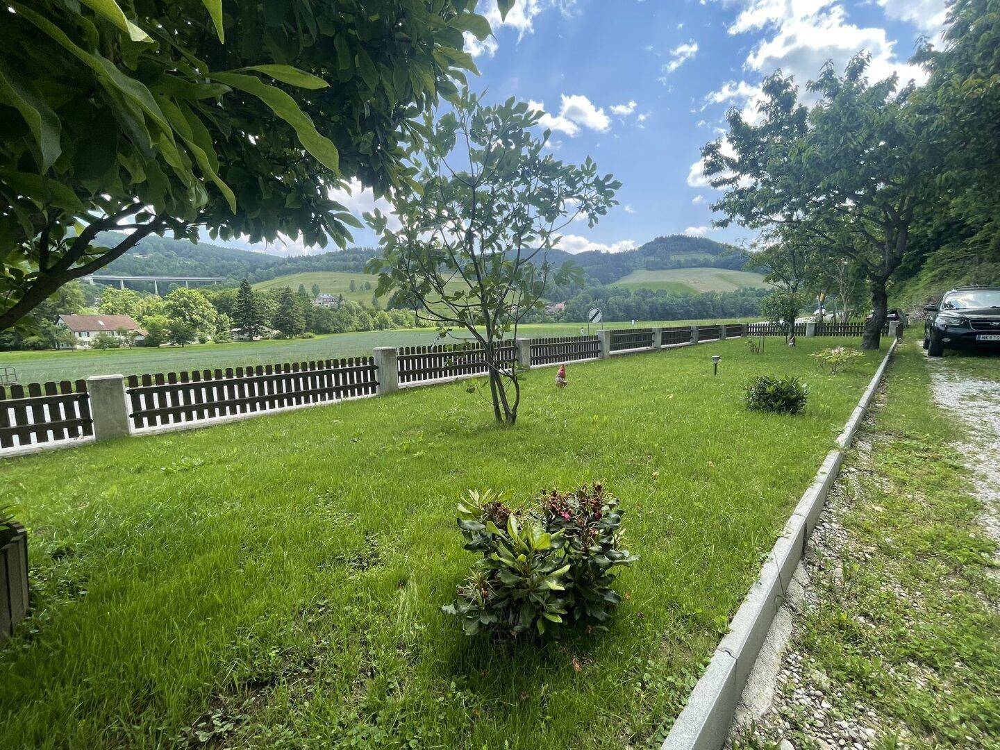 Gepflegter Gartenbereich mit Rasenfläche und Holzzaun, umgeben von grüner Landschaft und Hügeln.