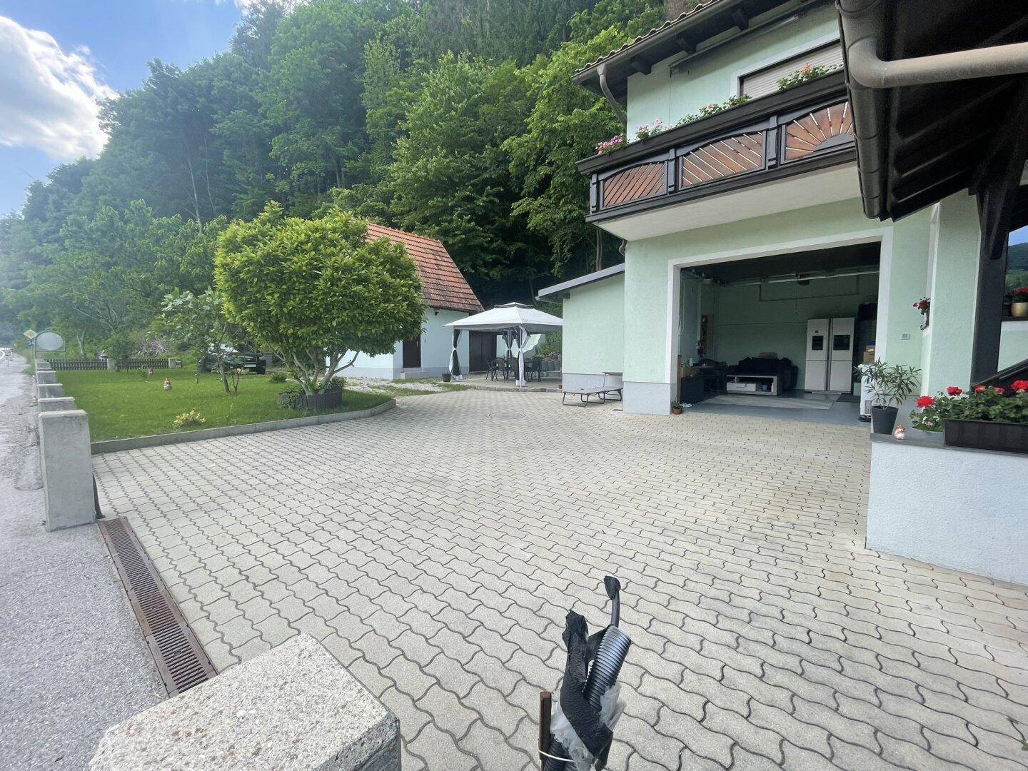 Vorderansicht des Hauses mit gepflastertem Hof, offener Garage und umgebender grüner Hügellandschaft.