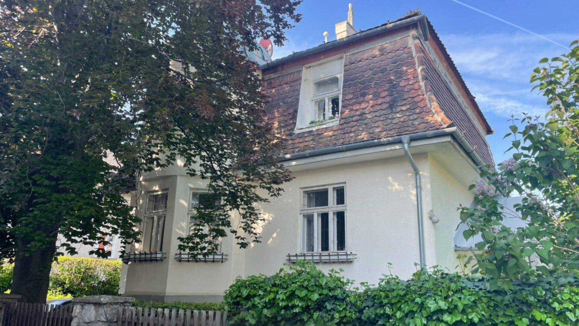 Charmantes Haus mit Mansarddach, umgeben von Bäumen und grüner Bepflanzung.