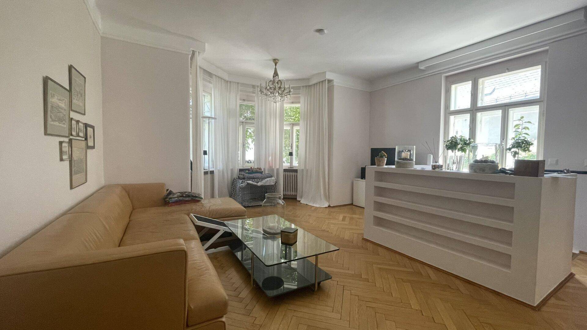 Helles Wohnzimmer mit Fischgrätparkett, beiger Couch und großen Fenstern mit viel Tageslicht.