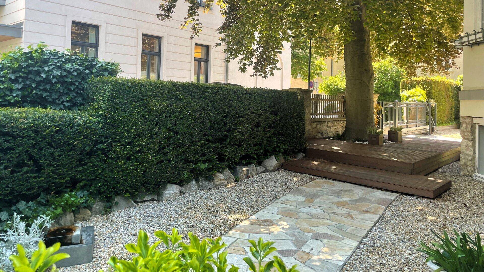 Gepflegter Gartenbereich mit Kiesweg, Hecke und einer Holzterrasse unter einem Baum.