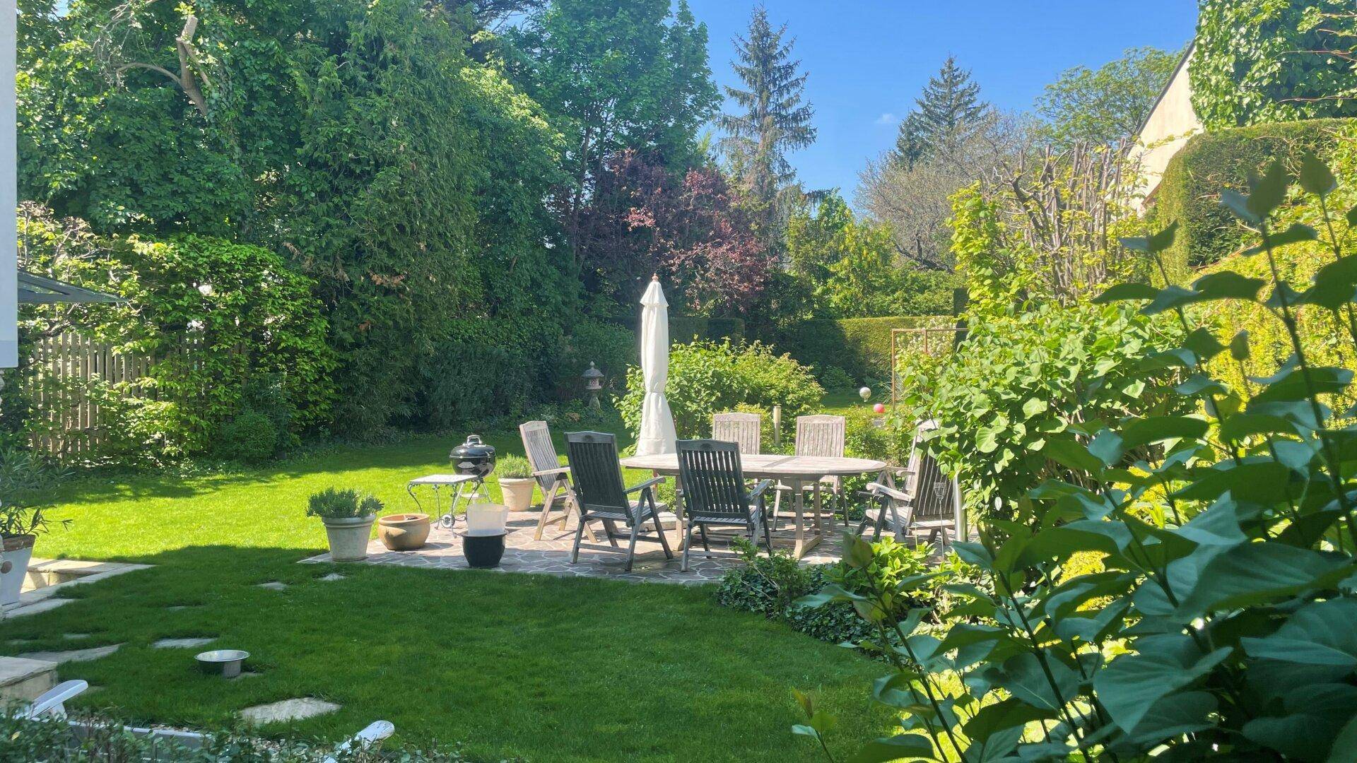 Weitläufiger Garten mit Rasenfläche, großer Steinterrasse und Gartenmöbeln unter Sonnenschirm.
