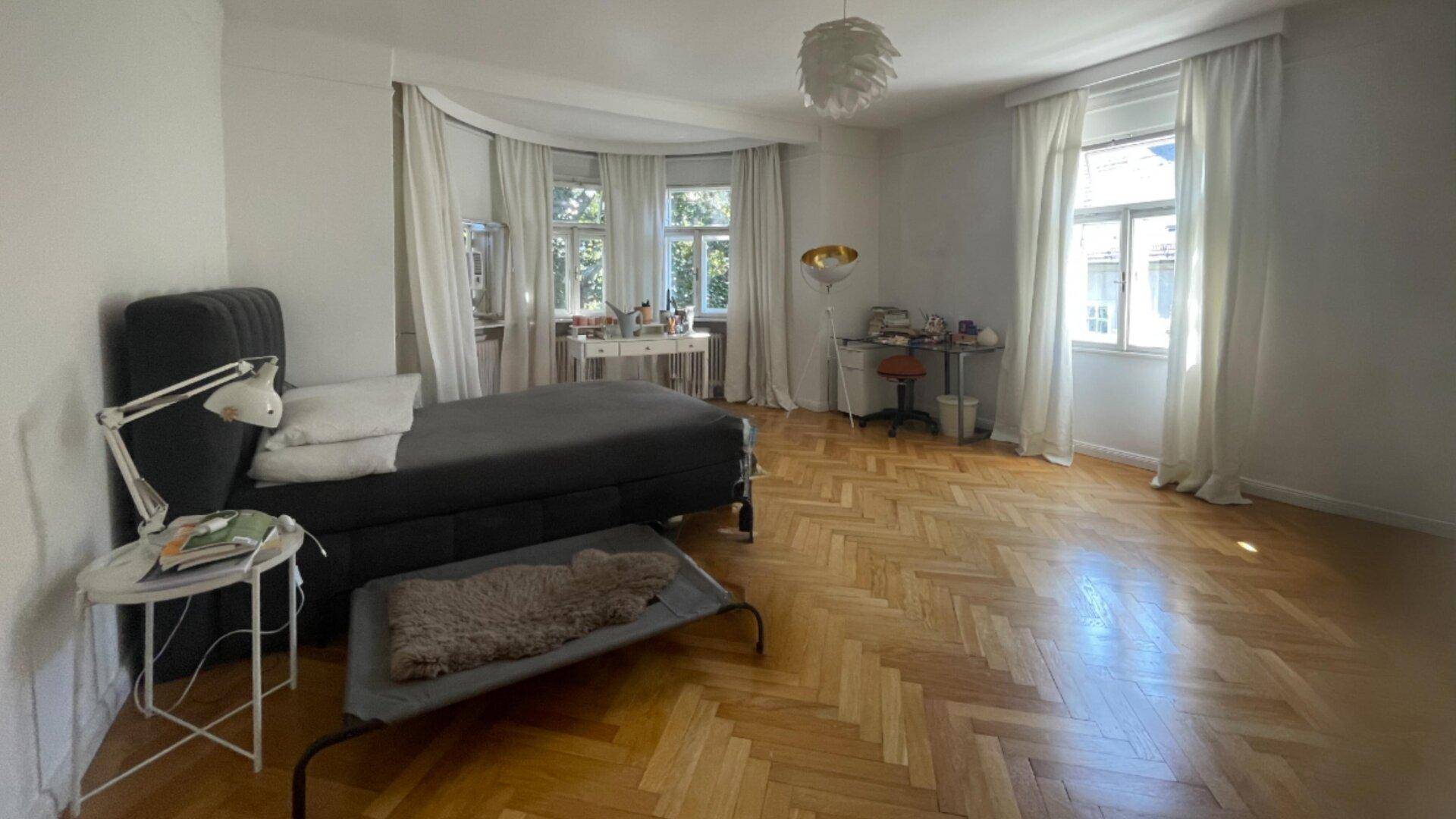Helles Schlafzimmer mit Fischgrätparkett, großem Fenster und minimalistischer Einrichtung.