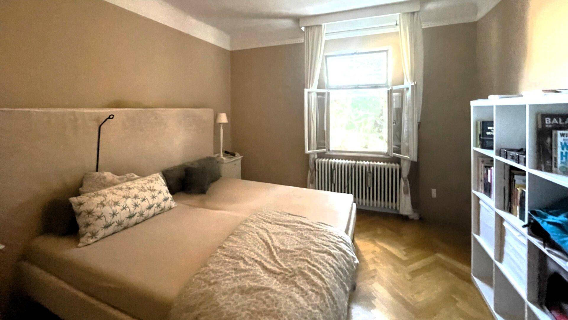 Gemütliches Schlafzimmer mit Doppelbett, Fischgrätparkett und einem Fenster mit Heizkörper.