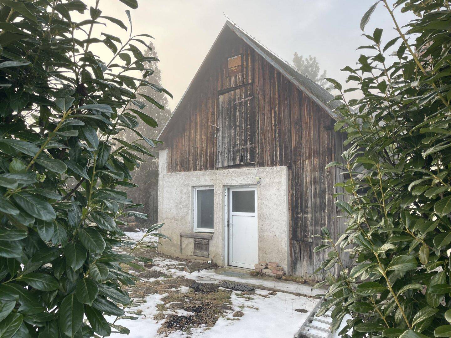 Rustikales Nebengebäude mit Holzwänden im oberen Bereich und verputztem Sockel, umgeben von Büschen und Schnee.
