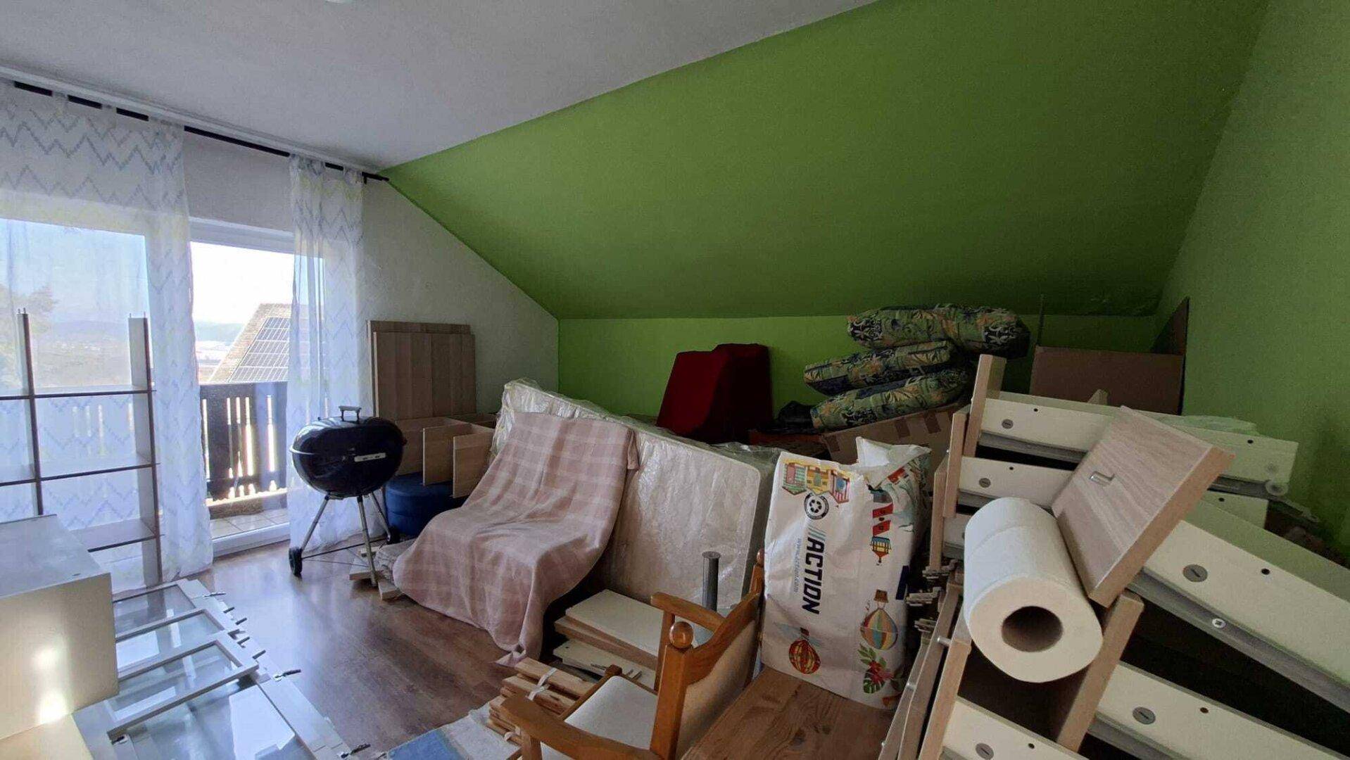 Dachzimmer mit schräger Decke, Balkonzugang und gelagerten Gegenständen.