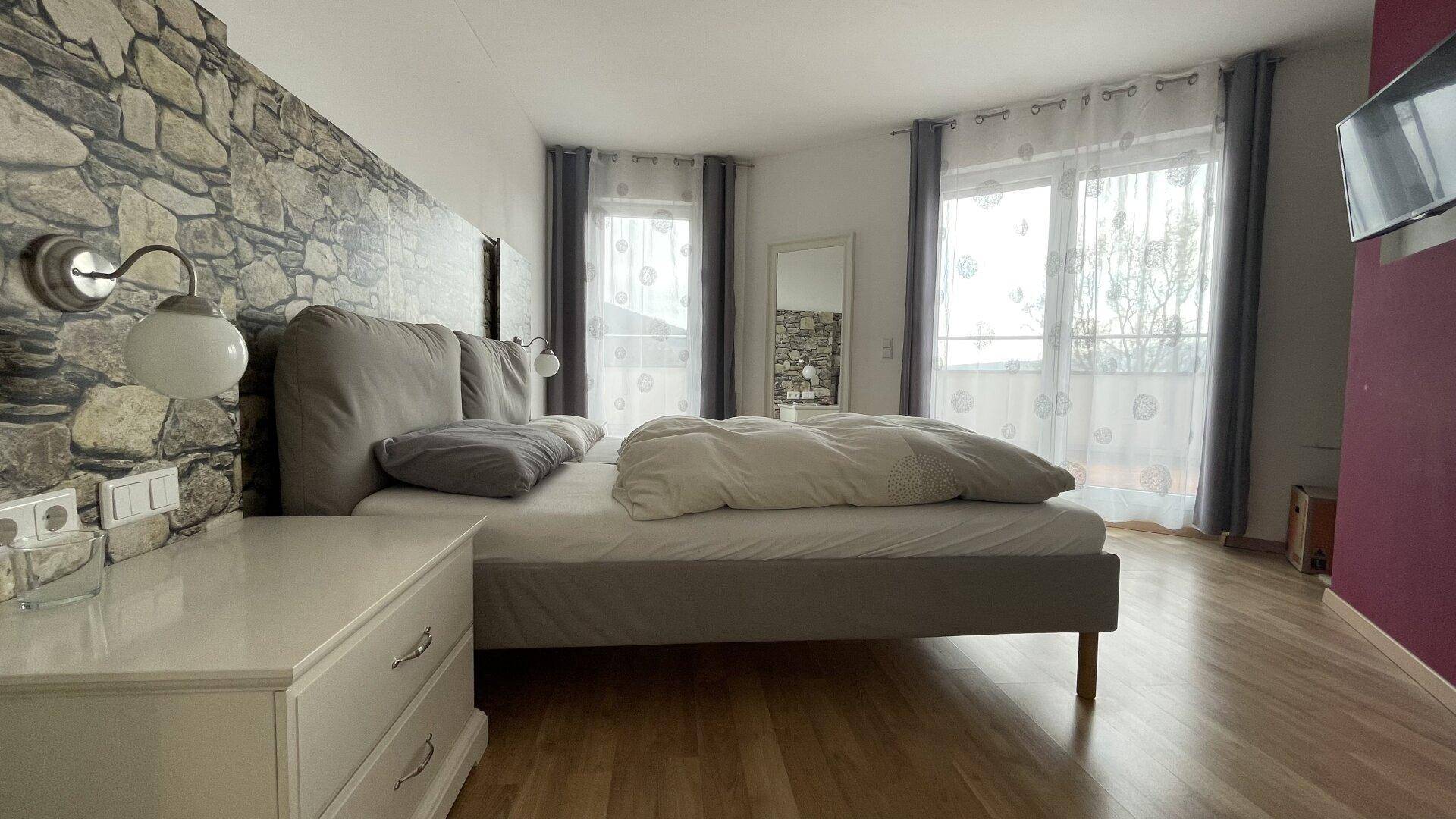Schlafzimmer mit Doppelbett, Steinwandakzent und Zugang zu einem französischen Balkon.
