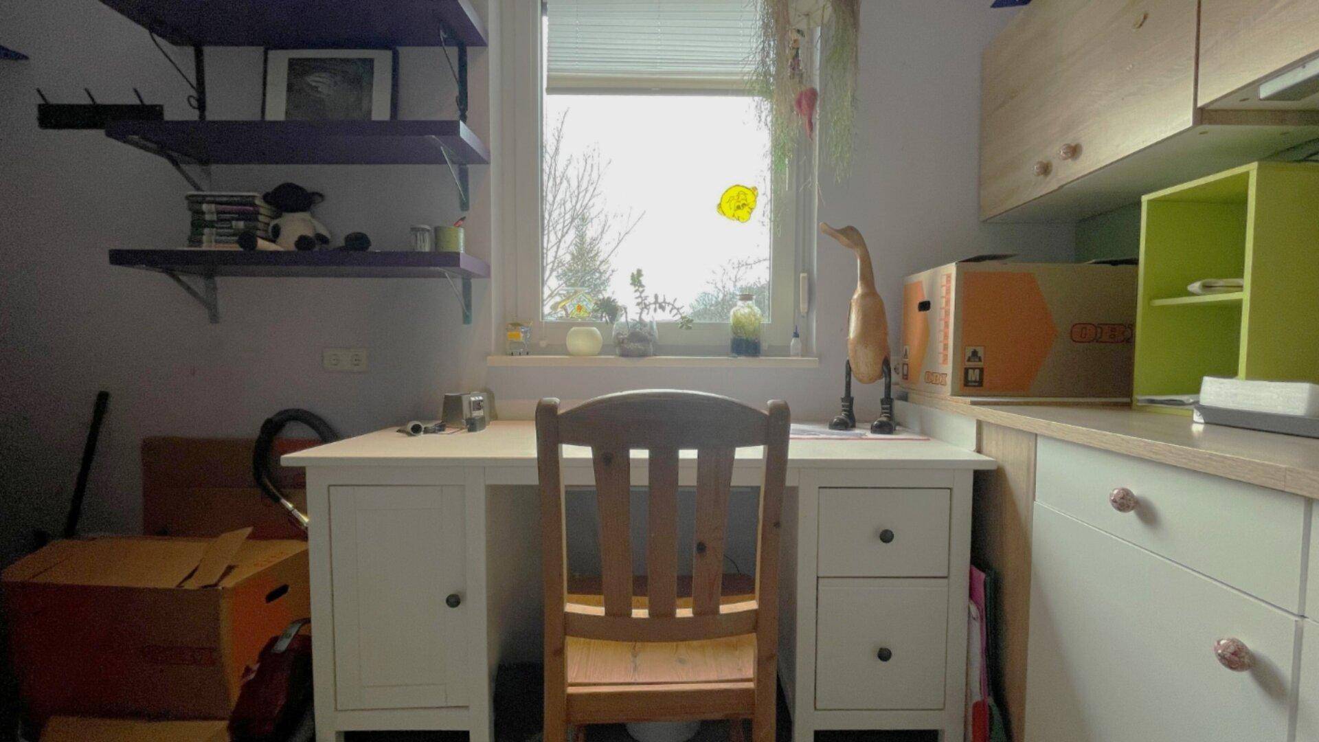 Kleines Arbeitszimmer mit Schreibtisch, Regal und Fenster mit Blick ins Grüne.