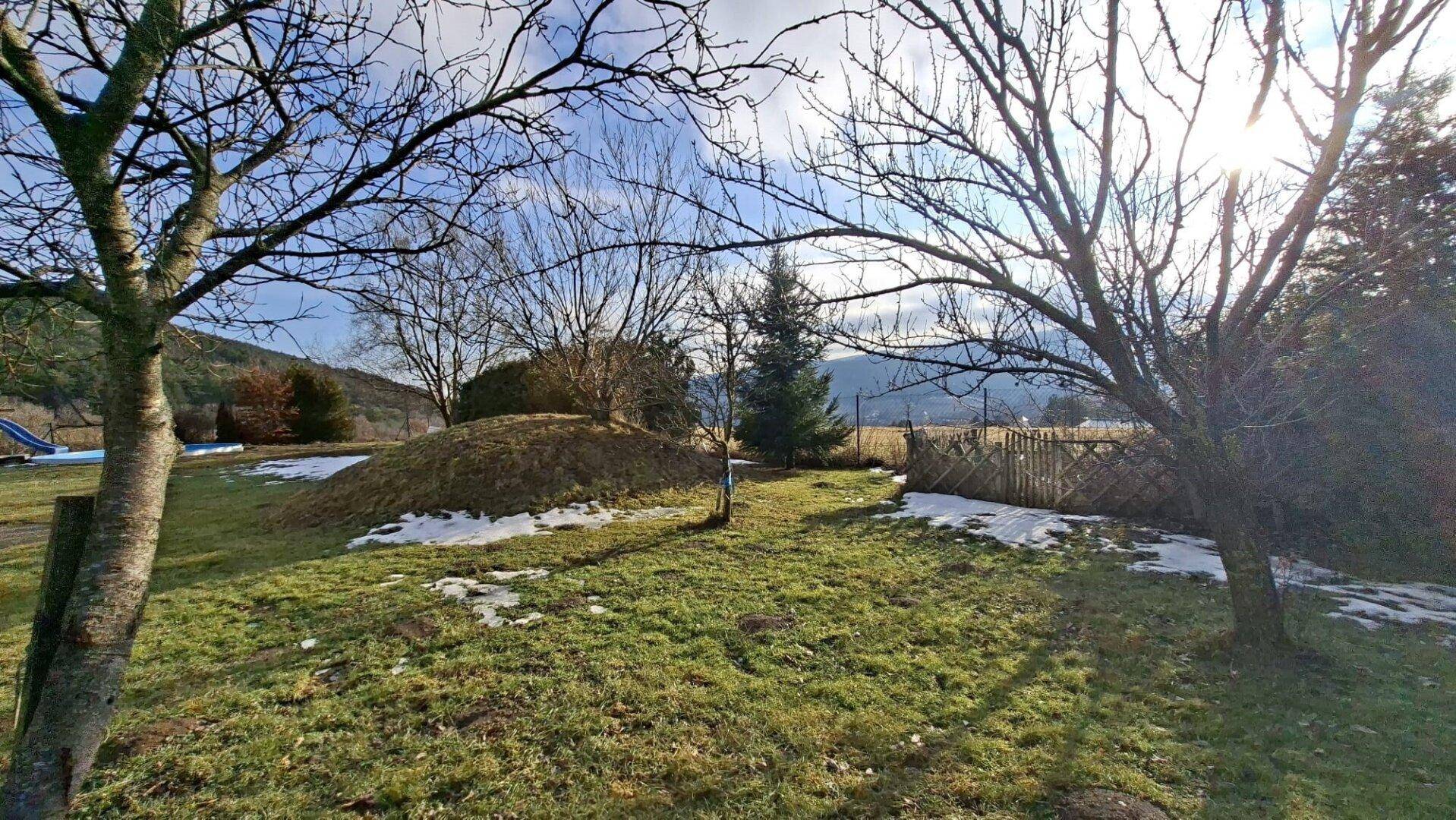 Sonniger Garten mit Bäumen, Rasenflächen und Resten von Schnee, umgeben von natürlicher Landschaft.