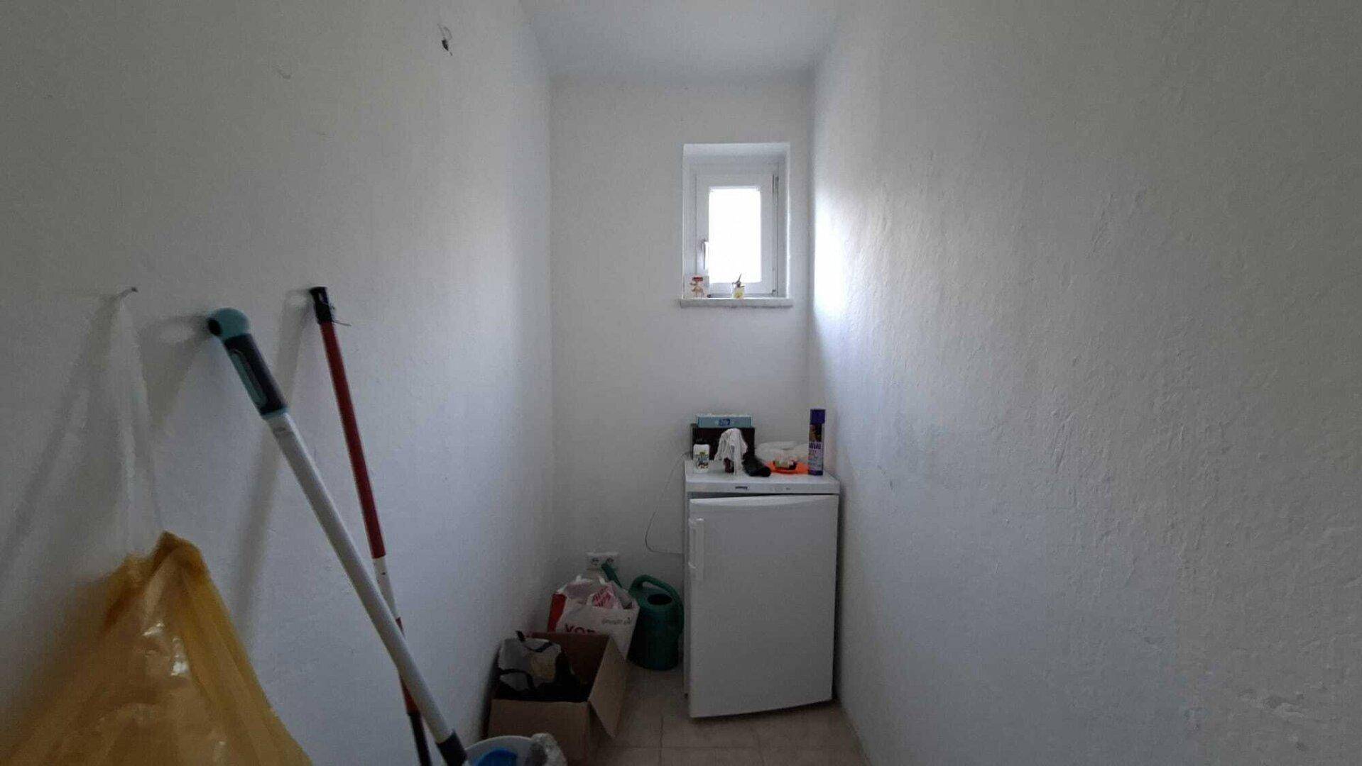 Kleiner Abstellraum mit Fenster, Fliesenboden und einem Kühlschrank.