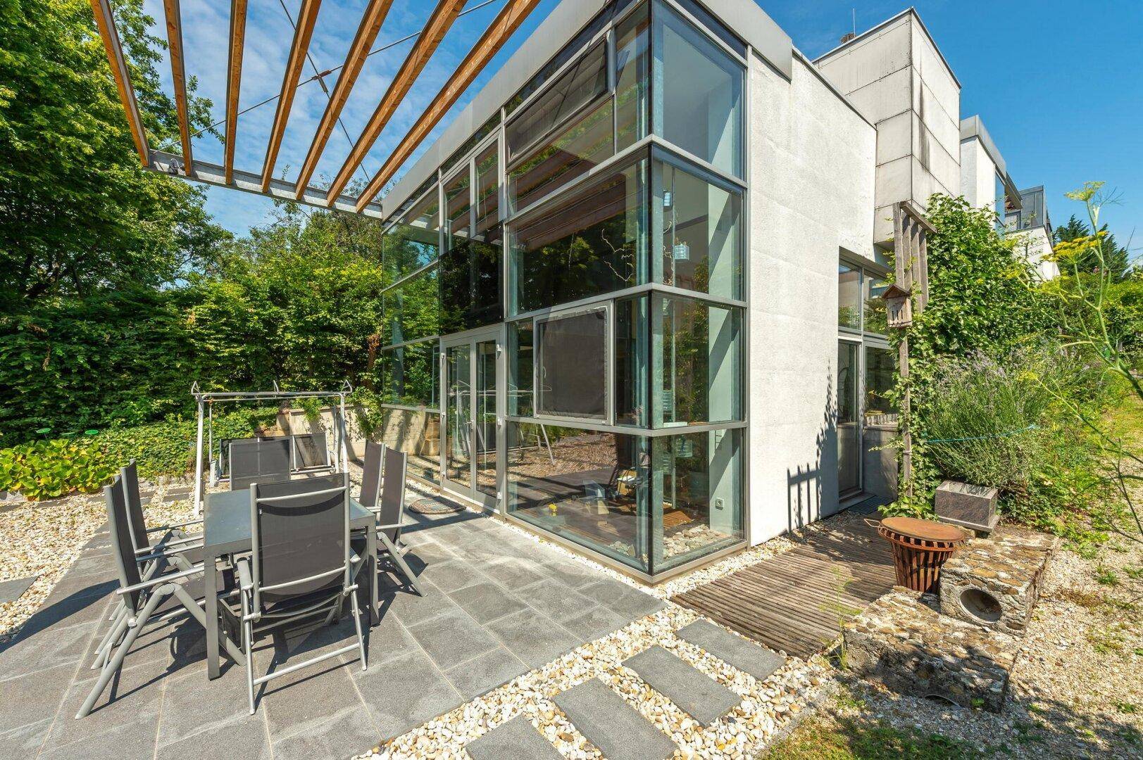 Sonniger Außenbereich mit Terrasse, Gartenmöbeln und moderner Glasfassade des Hauses.