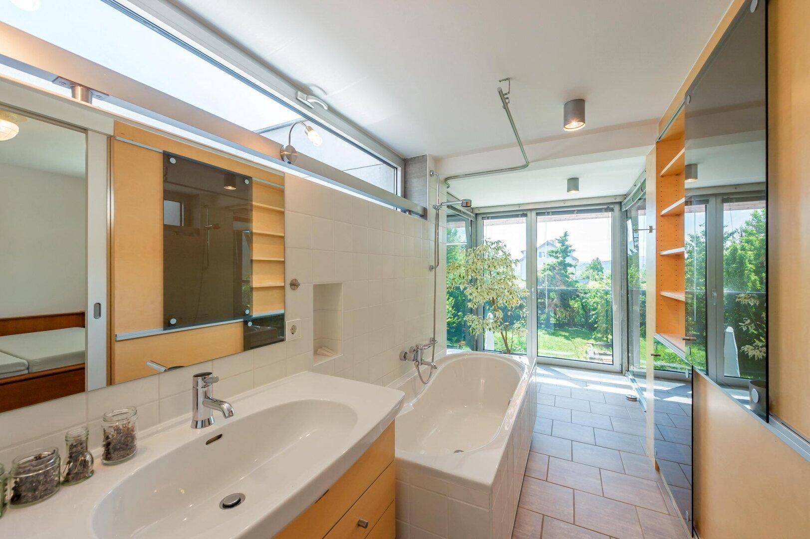 Geräumiges Badezimmer mit Badewanne, Doppelwaschbecken und großen Fenstern mit Gartenblick.