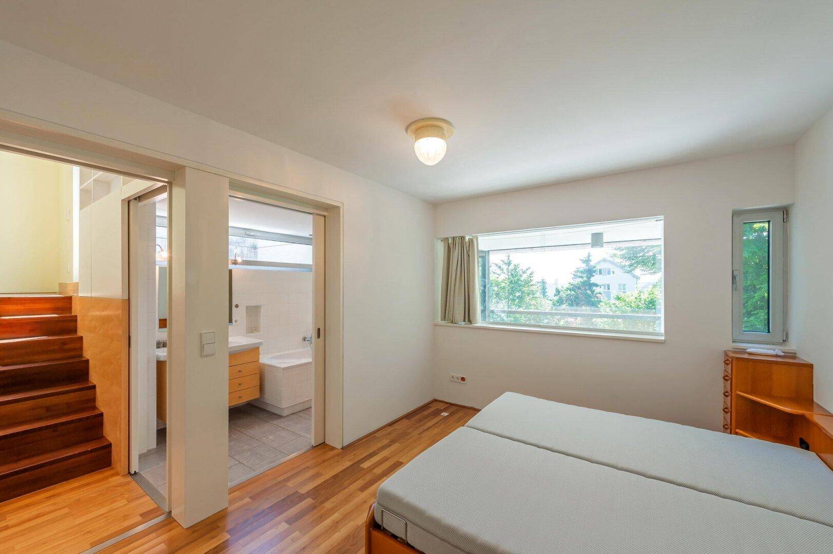 Schlafzimmer mit Holzboden und Zugang zu einem hellen Badezimmer mit Badewanne.