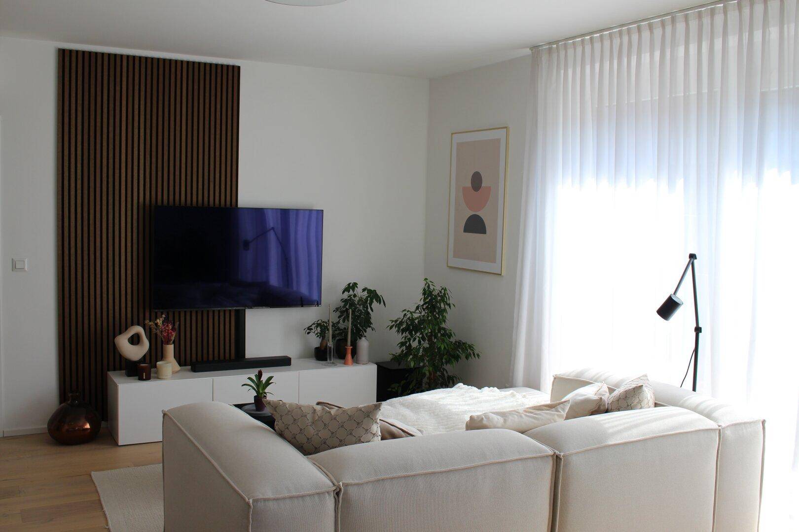 Modernes Wohnzimmer mit großem Sofa, Fernseher und bodentiefen Fenstern für viel Tageslicht.
