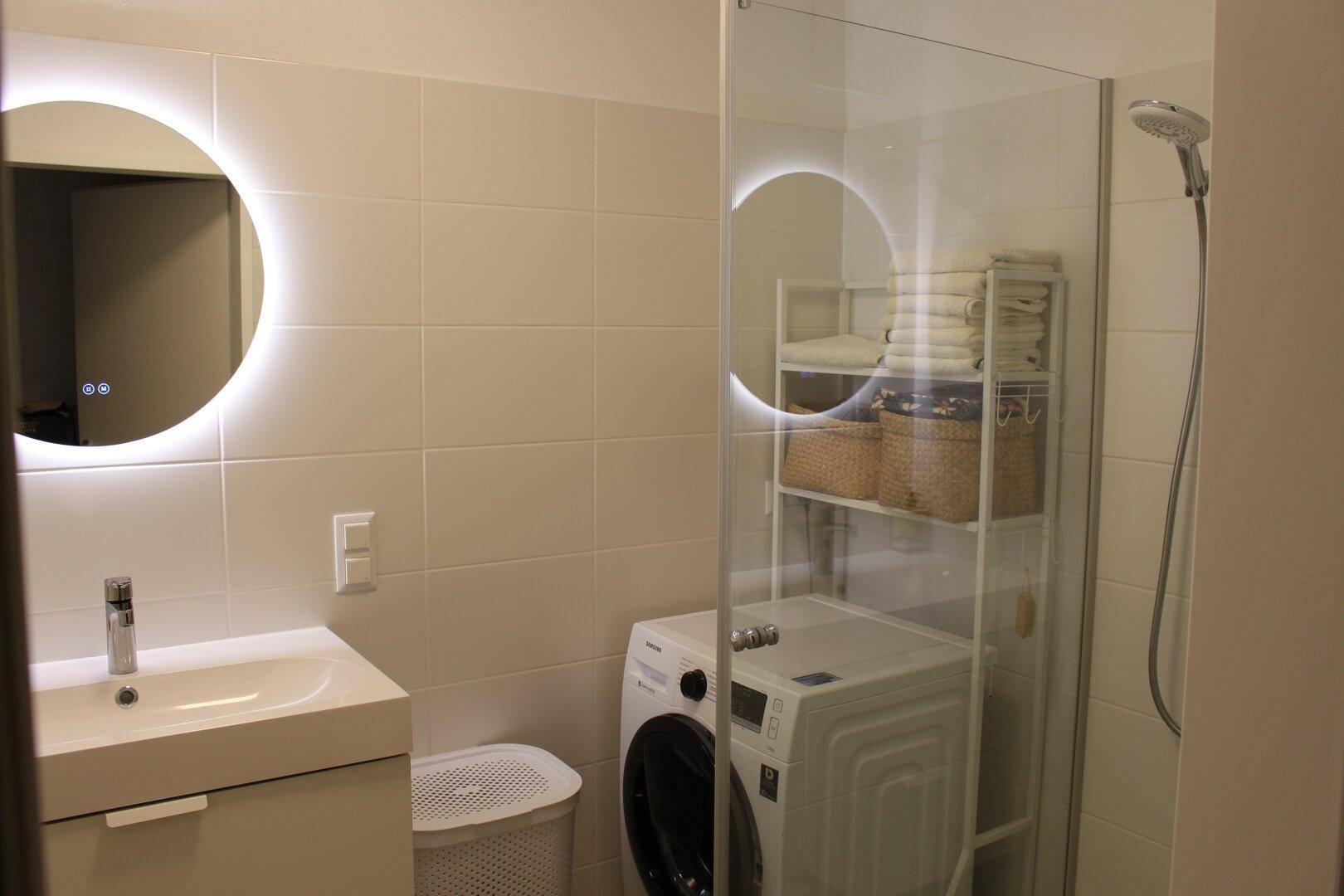 Modernes Badezimmer mit Dusche, Waschmaschine und beleuchtetem Spiegel, praktisch und hell gestaltet.