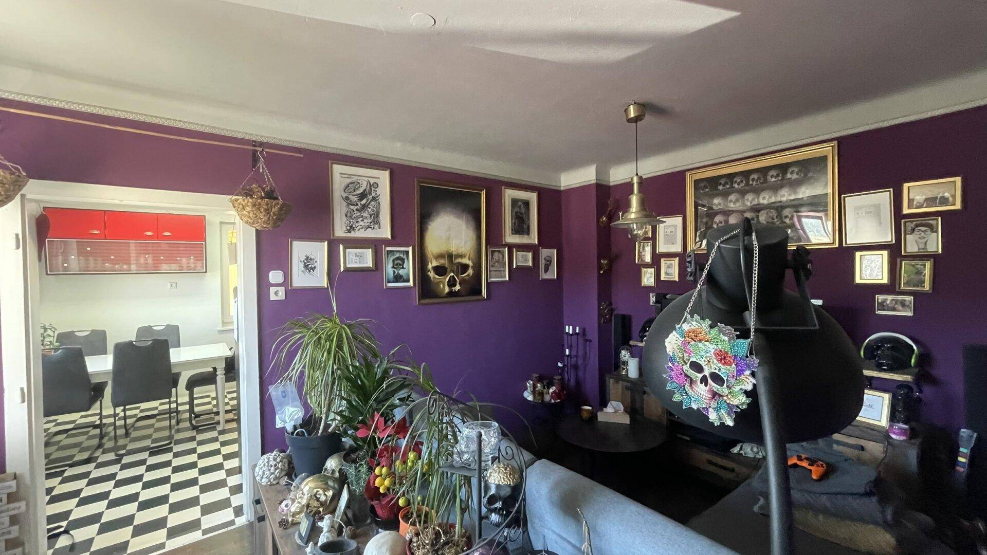 Wohnzimmer mit lila Wänden, vielen Bildern und Blick in die Küche.