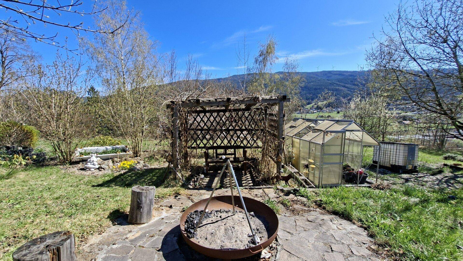 Gartenbereich mit Feuerstelle, Pergola und einem kleinen Gewächshaus.