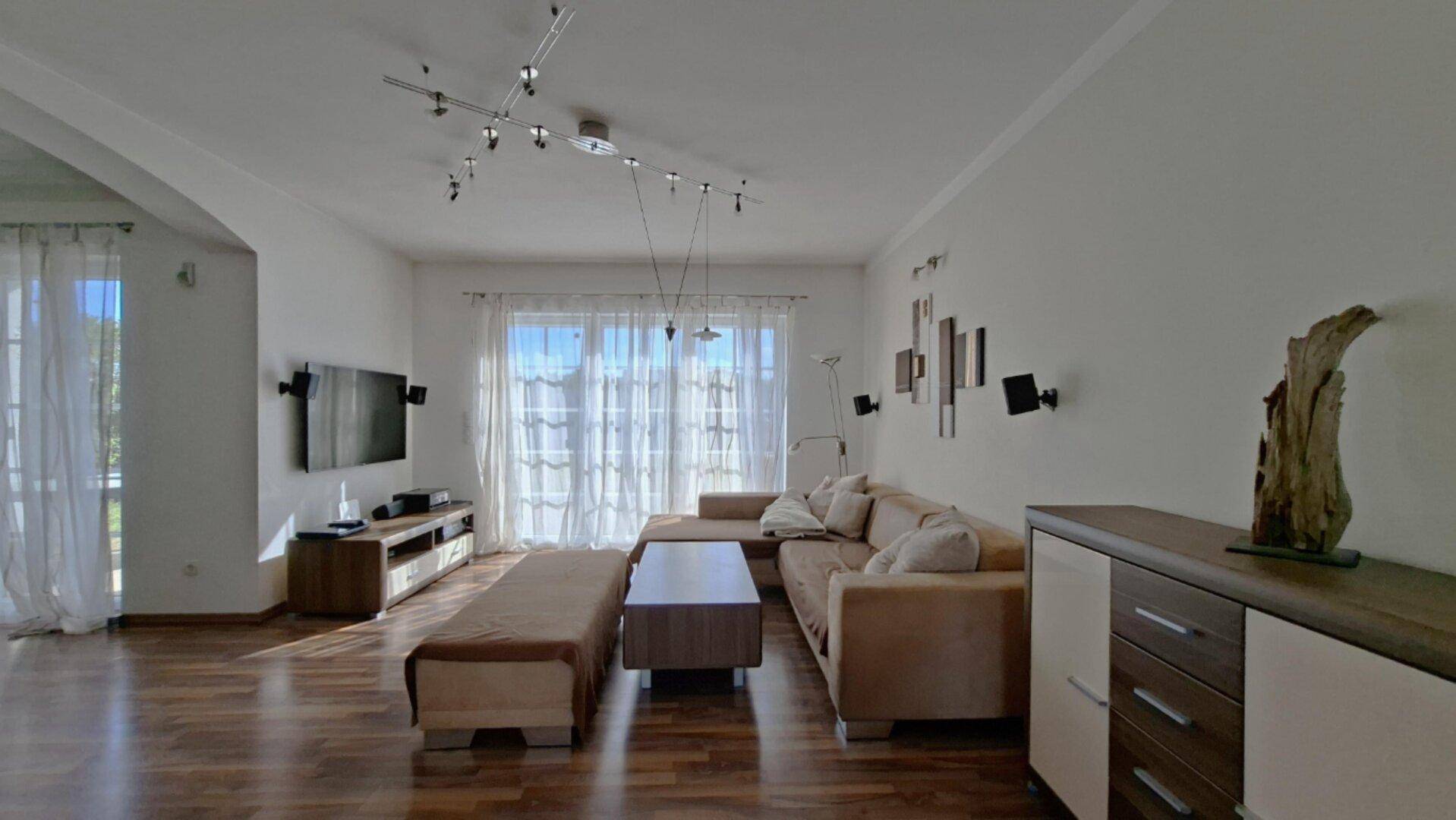 Helles Wohnzimmer mit beigem Sofa, Holzboden und großen Fenstern für viel Tageslicht.