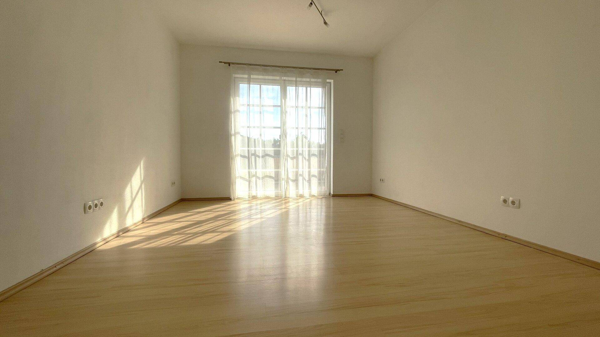 Leerer Raum mit hellem Holzboden und einer großen Glastür, die viel Sonnenlicht hereinlässt.