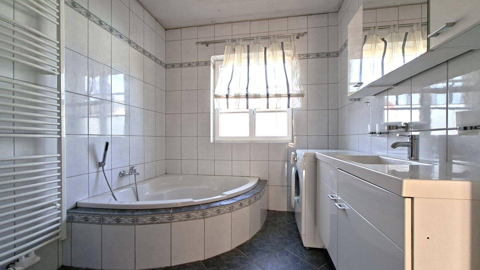 Gefliestes Badezimmer mit Eckbadewanne, Waschbecken und Fenster für natürliche Belüftung.