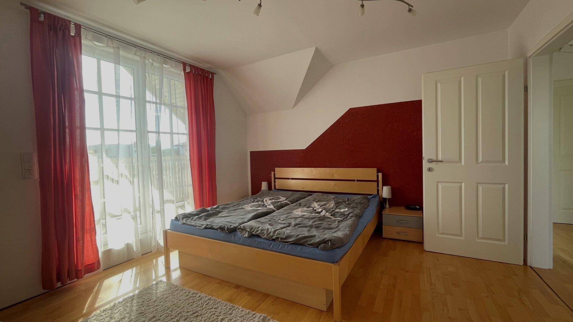 Gemütliches Schlafzimmer mit Doppelbett, roter Akzentwand und großen Fenstern mit Vorhängen.