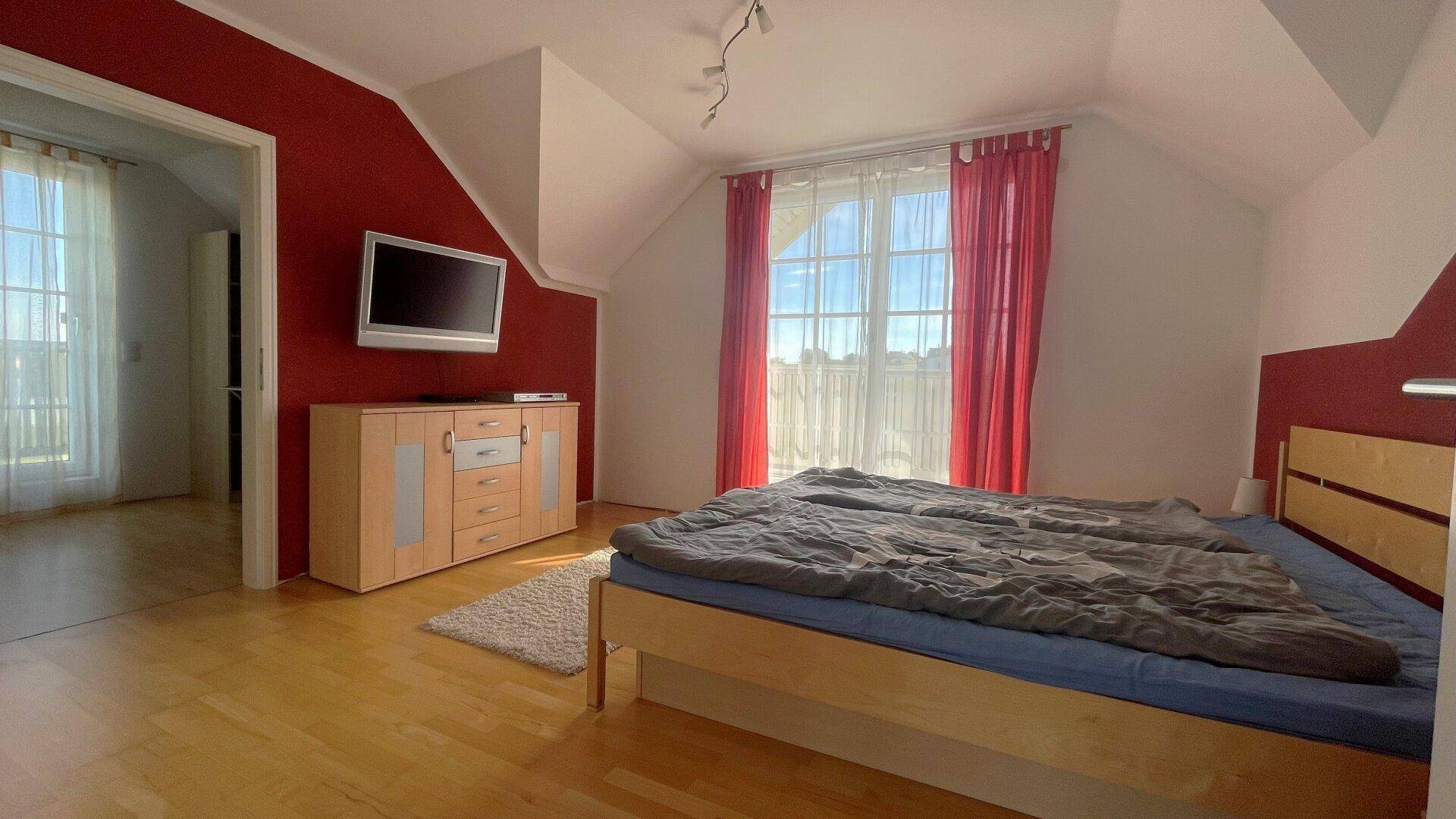 Schlafzimmer mit Fernseher auf einer Kommode, roter Wand und Zugang zu einem weiteren Raum.