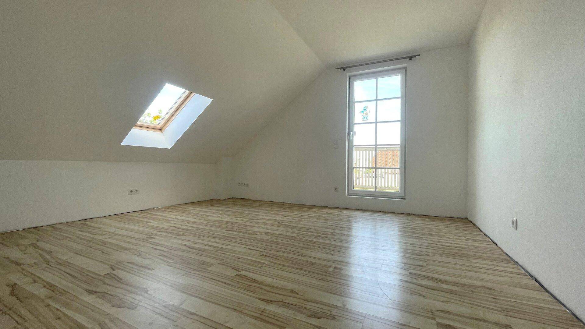 Heller, leerer Raum mit Holzboden, großem Fenster und Dachfenster in der Schräge.