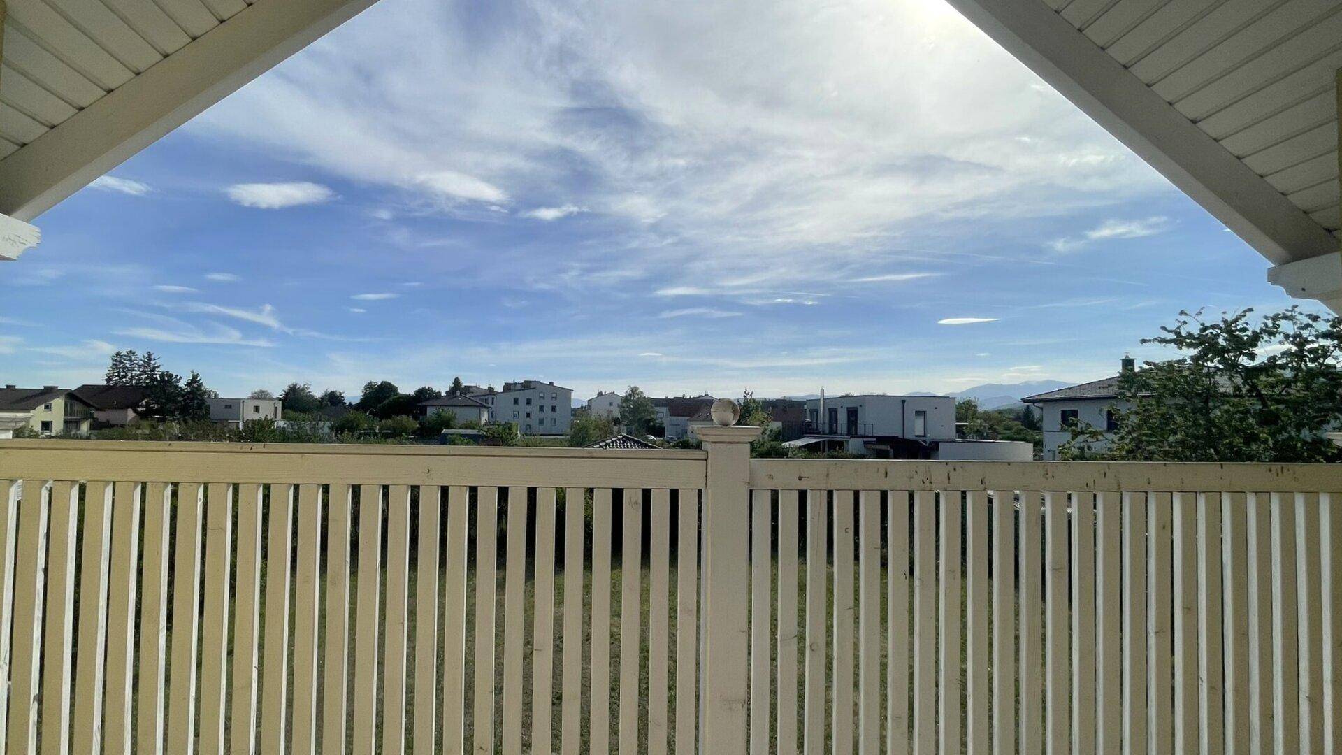 Ausblick von einem Balkon mit Holzgeländer auf die umliegende Wohngegend und Berge.
