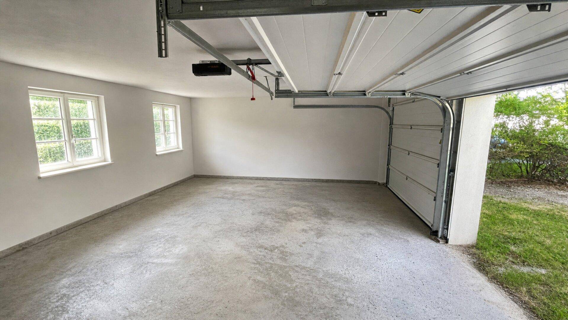 Geräumige, leere Garage mit Betonboden, zwei Fenstern und offenem Garagentor.