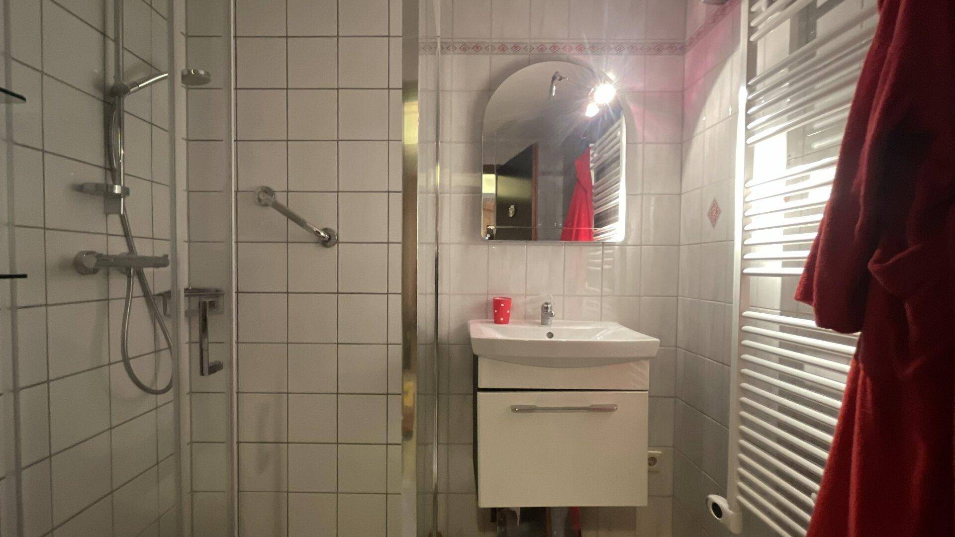 Helles Badezimmer mit weiß gefliesten Wänden, Dusche, Waschbecken und Handtuchheizkörper.