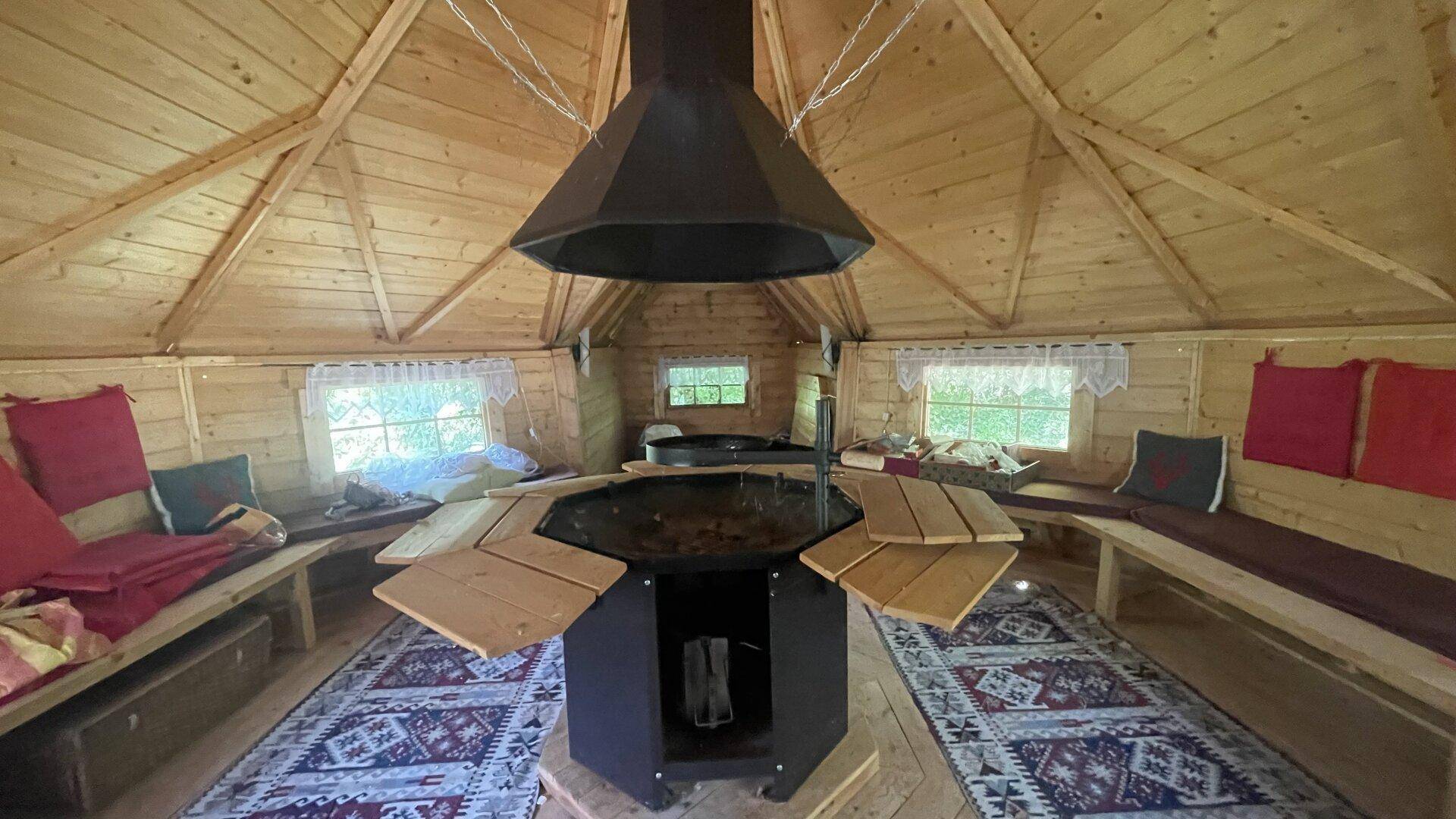 Gemütlicher Innenraum einer Holzhütte mit zentraler Grillstelle, Sitzbänken und rustikalem Charme.