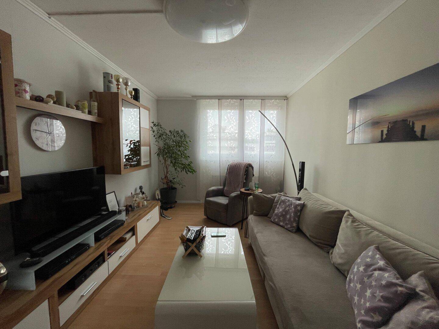 Helles Wohnzimmer mit beigem Sofa, TV-Möbel und hellem Laminatboden, gut beleuchtet.