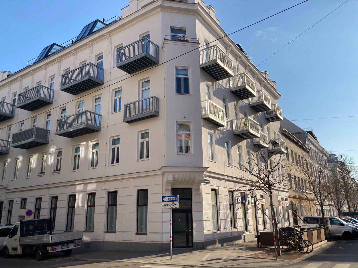 Eckgebäude mit klassischer Architektur, zahlreichen Balkonen und Geschäften im Erdgeschoss an einer belebten Straße.