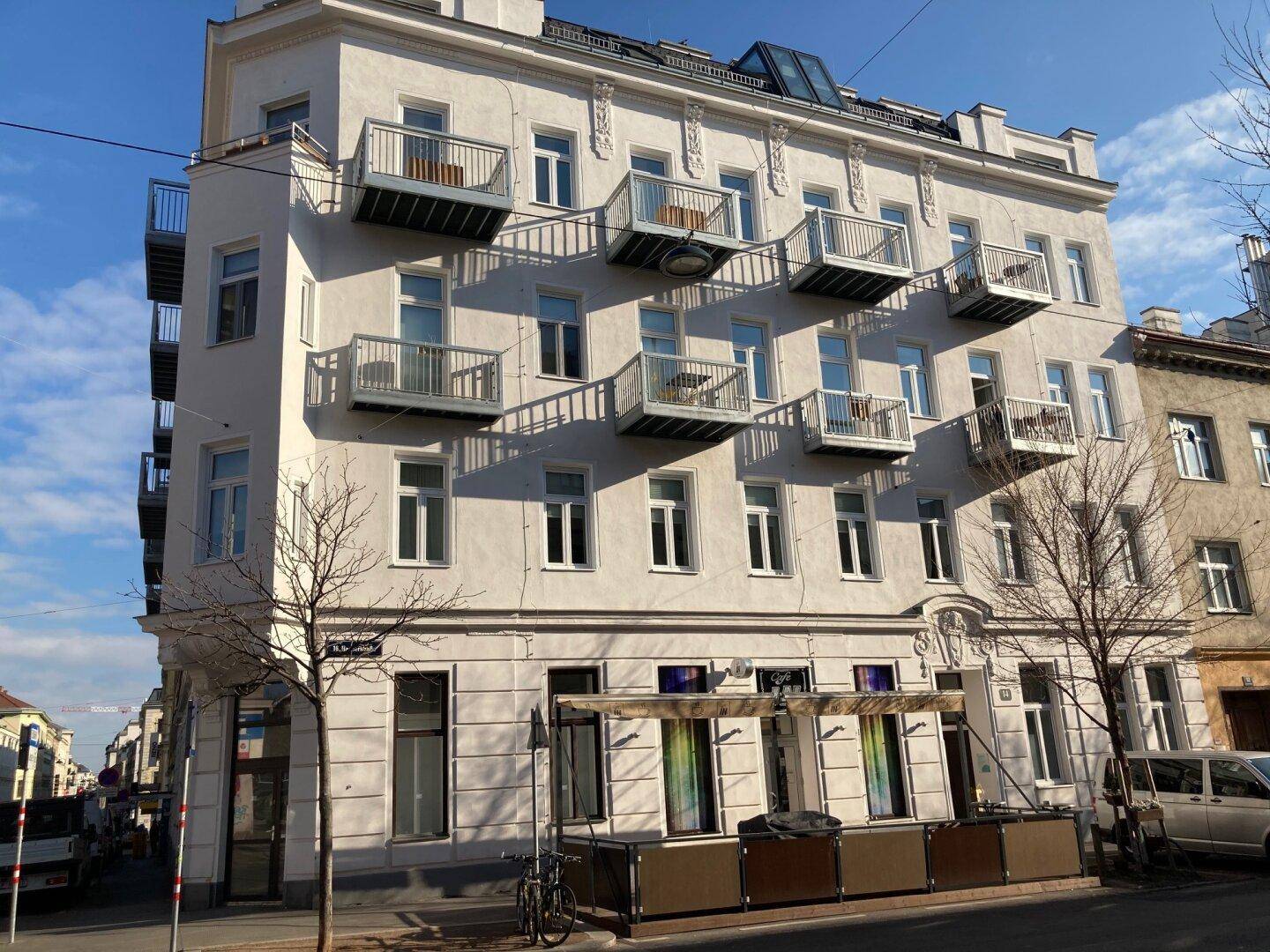 Weitwinkelansicht eines historischen Eckgebäudes mit vielen Balkonen und Geschäften im Erdgeschoss unter blauem Himmel.