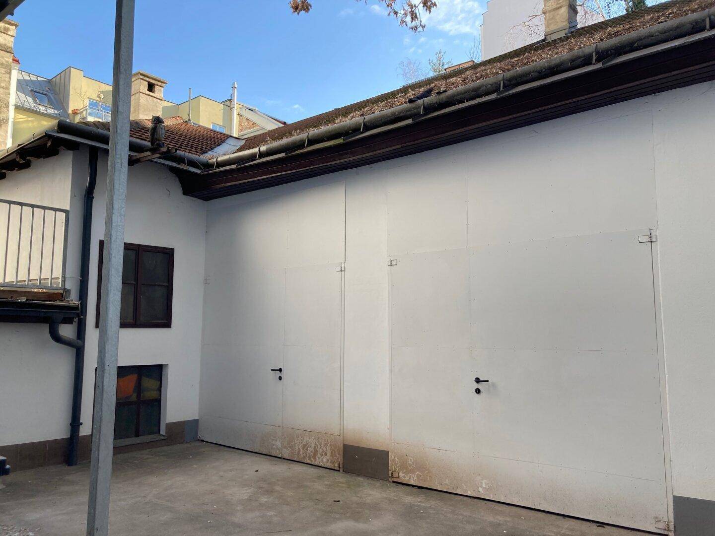 Rückseite eines Gebäudes mit zwei großen Toren und einem kleinen Fenster, umgeben von einer gepflasterten Fläche.