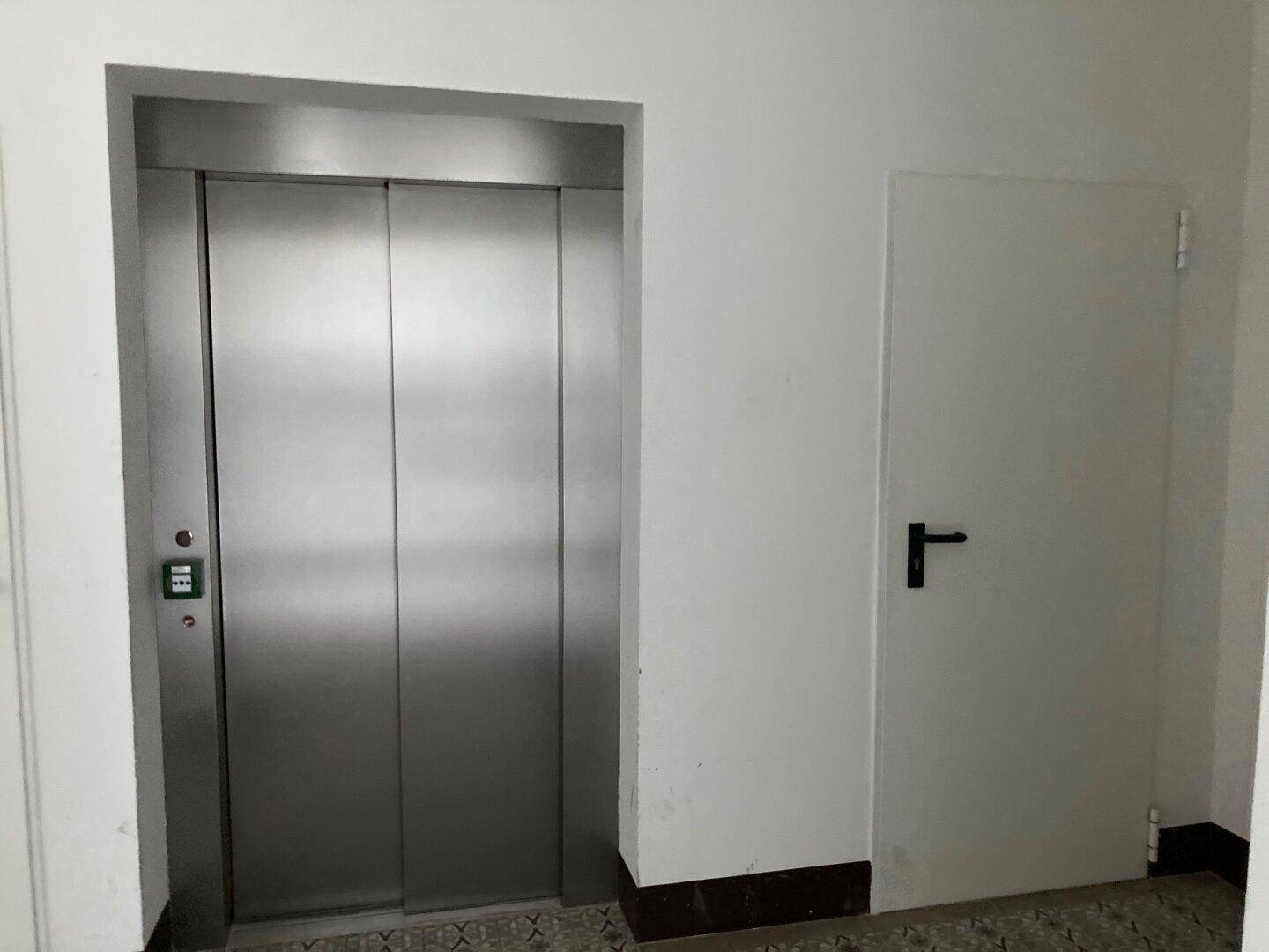 Moderner Aufzug mit Edelstahltüren in einem hellen Flur neben einer weißen Tür.