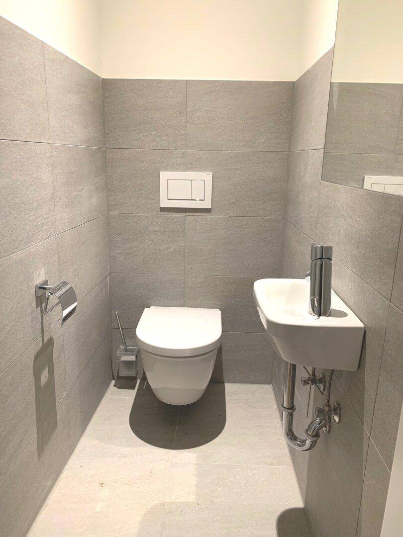 Modernes Gäste-WC mit grauen Fliesen, wandhängendem WC und kleinem Waschbecken.