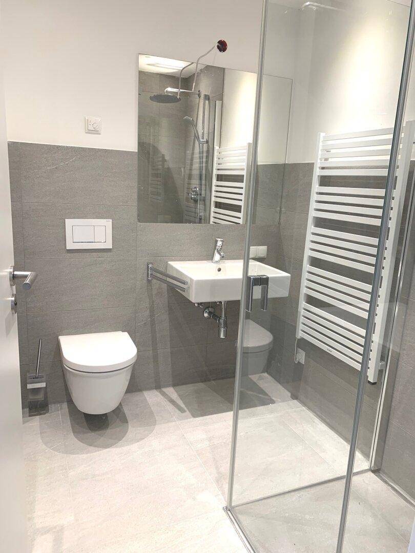 Modernes Badezimmer mit grauen Fliesen, Dusche, WC und beheiztem Handtuchhalter.