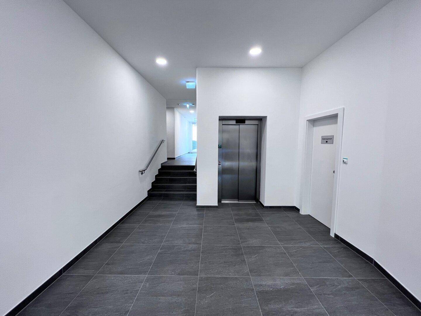 Heller Eingangsbereich mit Fliesenboden, Treppe und Aufzug.