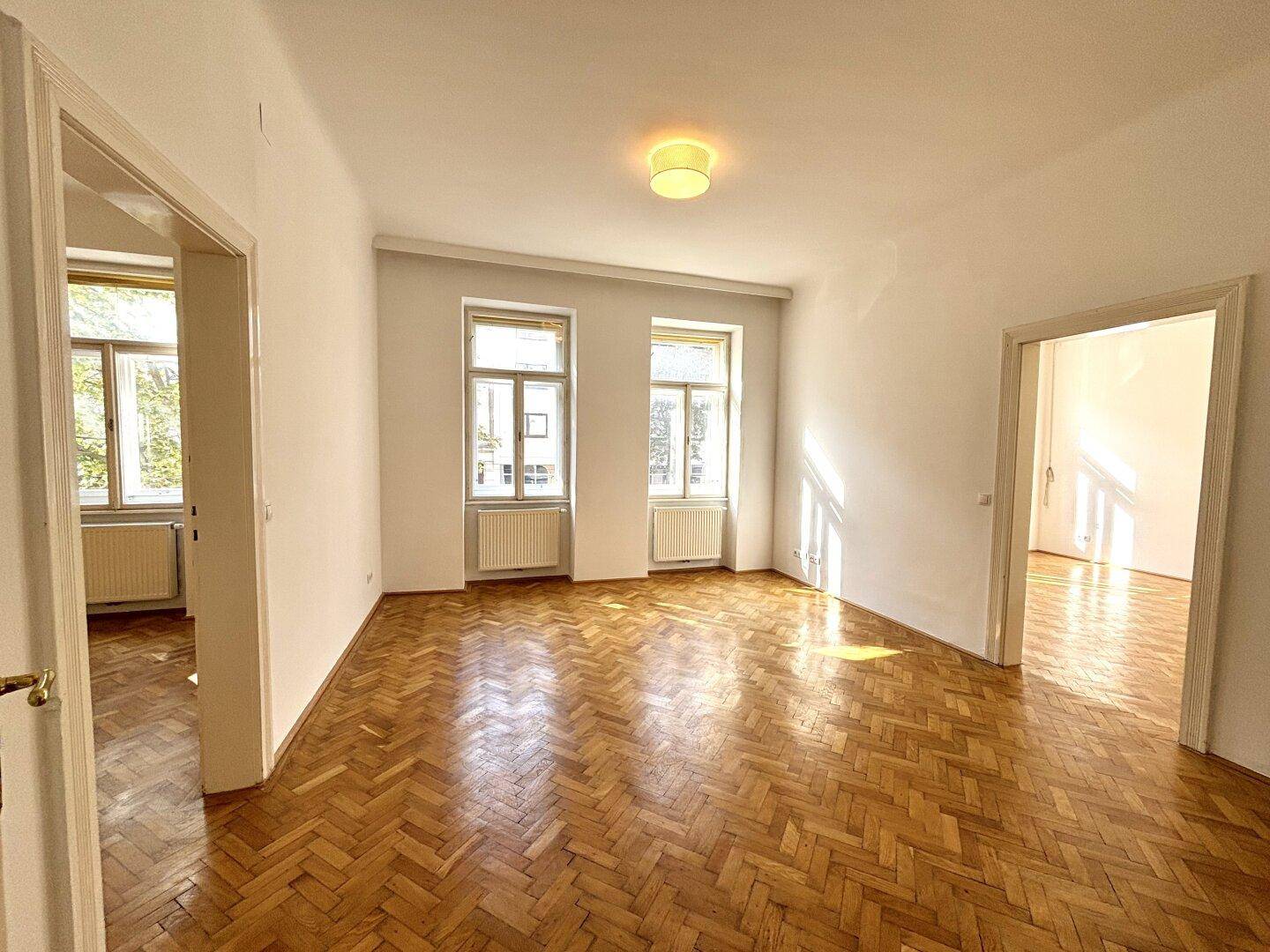 Geräumiges, leeres Wohnzimmer mit Fischgrätparkett und viel natürlichem Licht durch große Fenster.