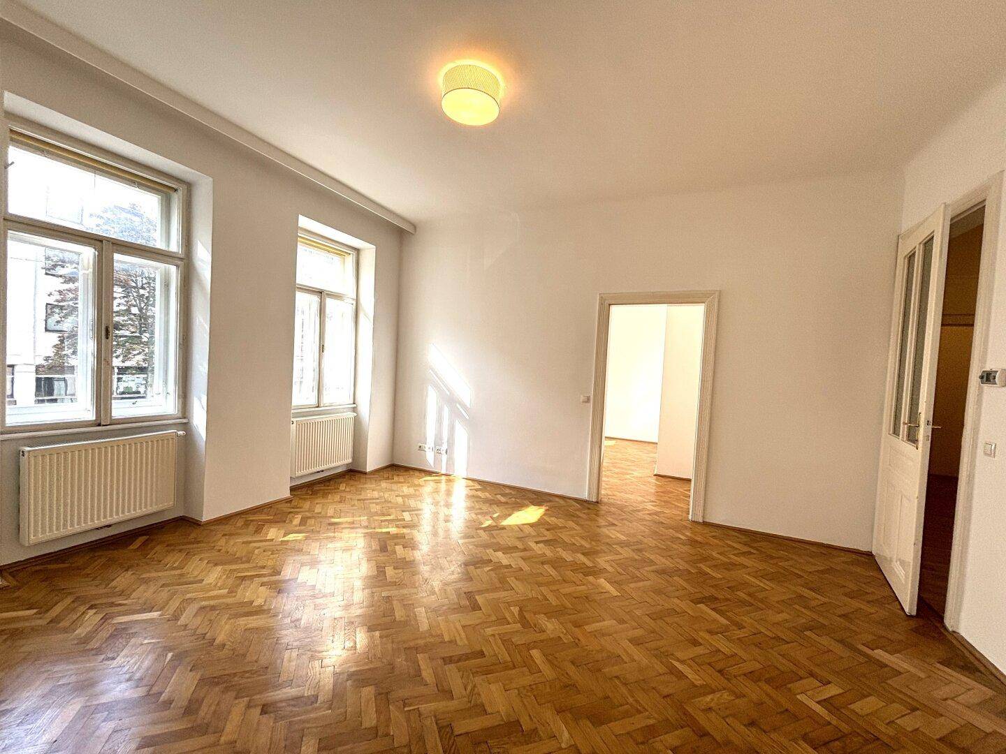 Helles, leeres Wohnzimmer mit Fischgrätparkett, hohen Decken und zwei großen Fenstern.