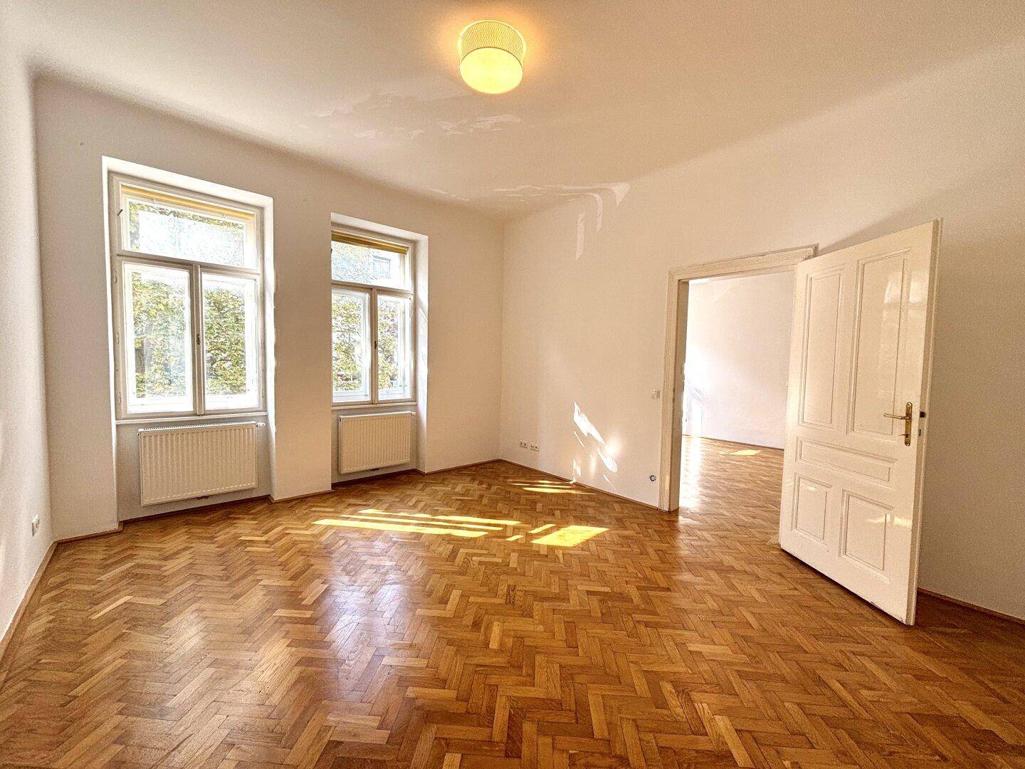 Großes, leeres Zimmer mit Fischgrätparkett und sonnigen Fenstern, bereit zur individuellen Gestaltung.